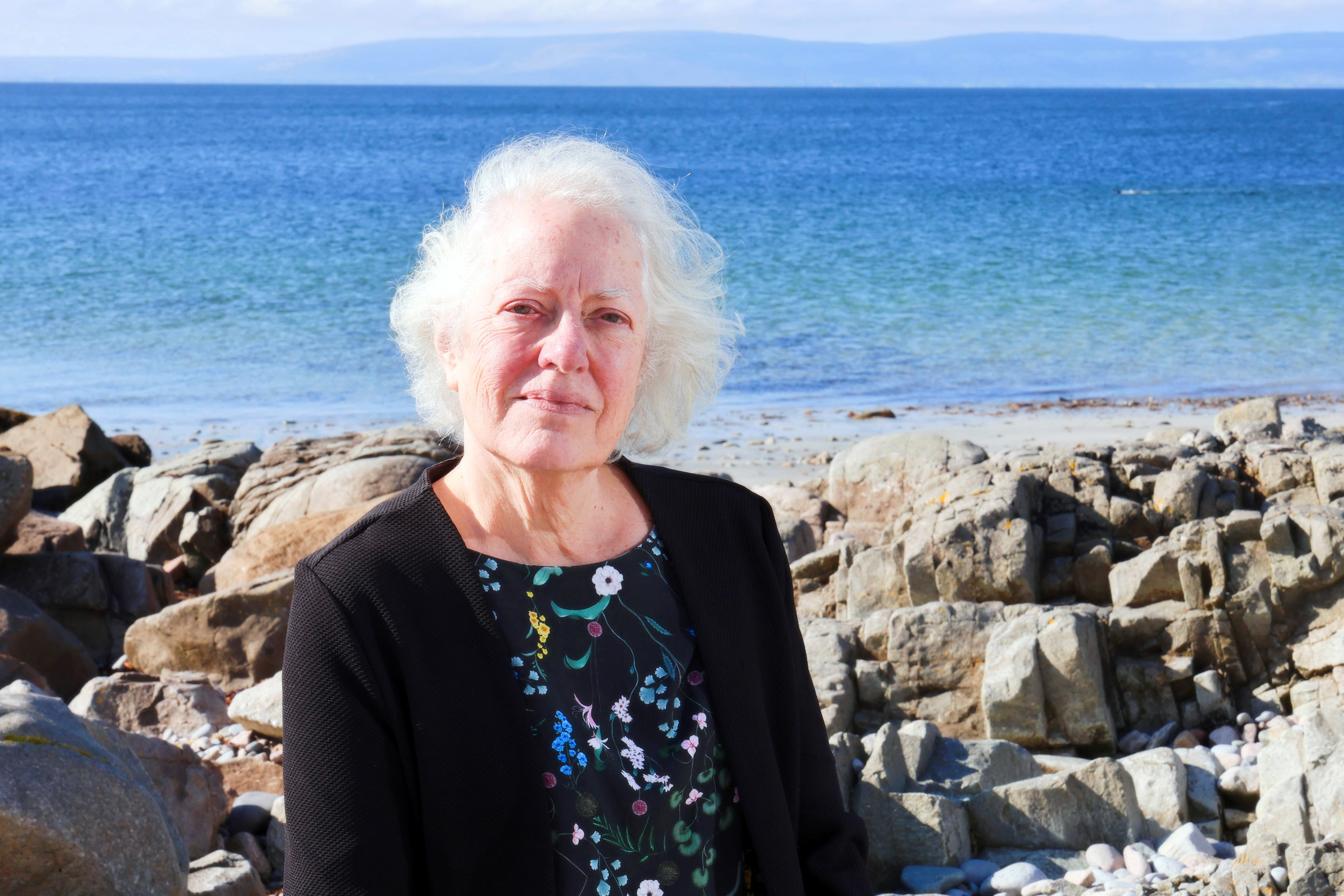 Jane O'Leary, composer, on a rocky beach, with the sea behind. The sun is shining. Photo Credit: Mike Shaughnessy